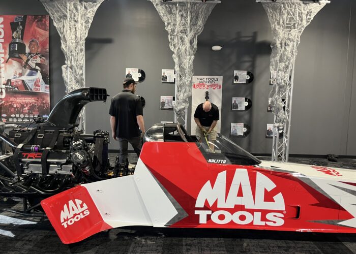Red and white race car in a showroom with MAC Tools branding and wall posters; people inspecting equipment in the background.