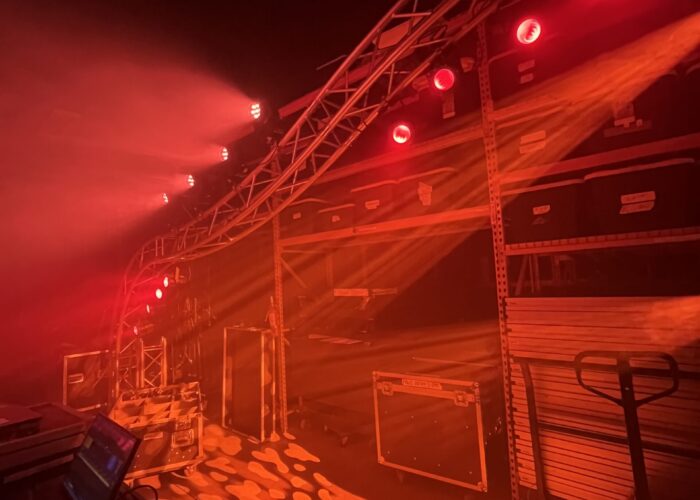 Backstage area at a concert with a red-lit truss, rigging, and equipment cases along a wall.