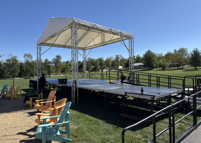 Outdoor outdoor stage with a white canopy supported by metal trusses, set on a grassy field with colorful Adirondack chairs in the foreground.